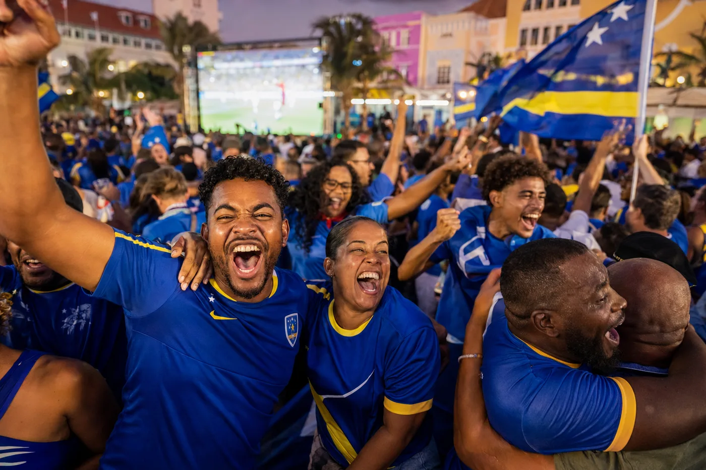 Juichende supporters in blauwe shirts met de vlag van Curaçao voor een groot scherm
